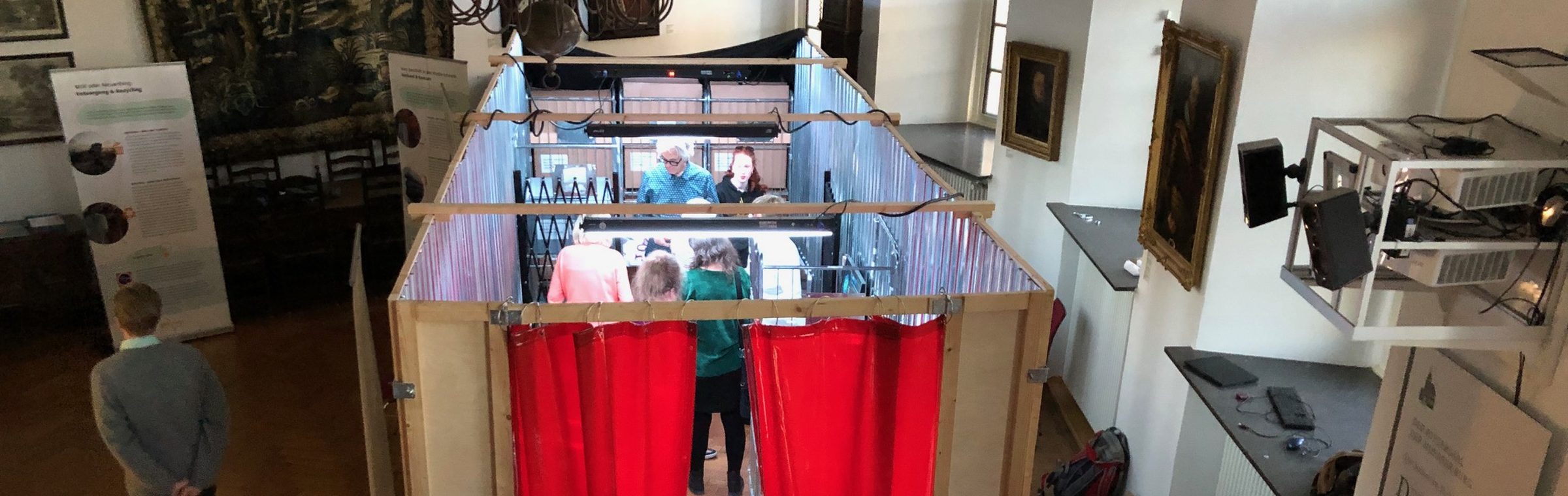 Blick von oben in den Container der Wahrheit: Besucherinnen und Besucher zwischen Gitterboxen und roten Vorhängen
